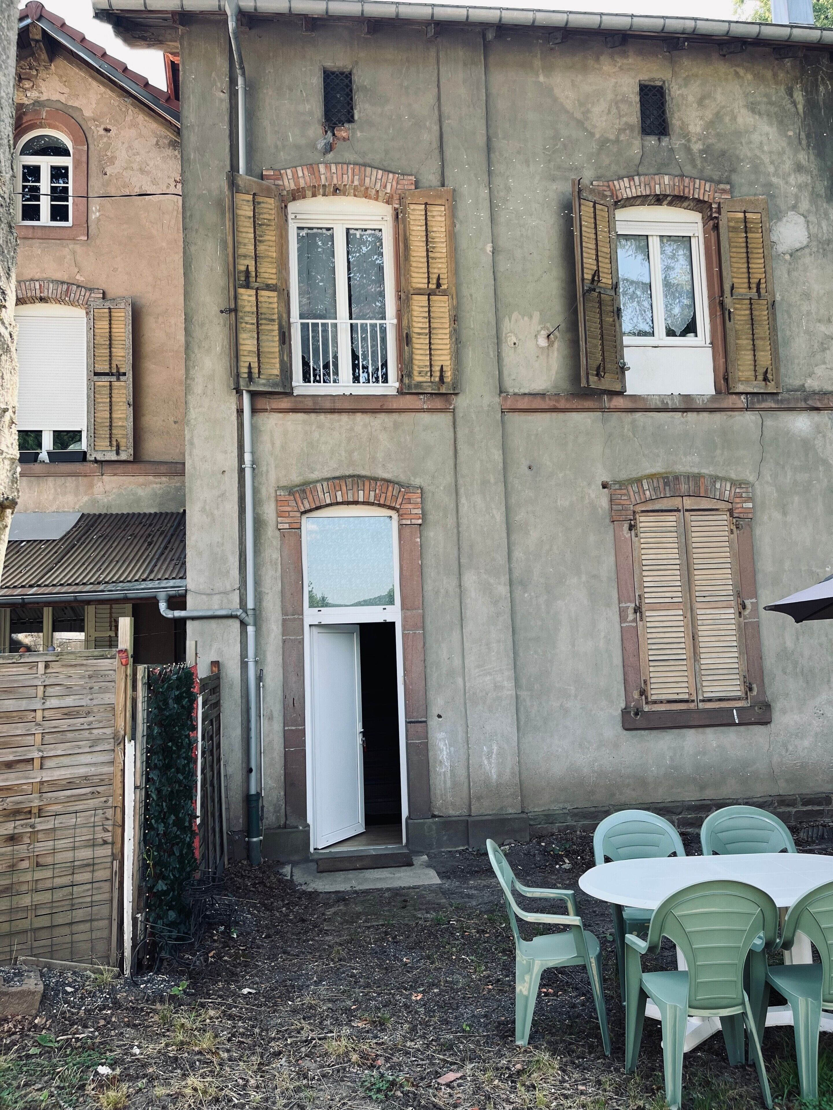 A building with multiple windows, some with shutters, a door, and a small garden area with plastic chairs.