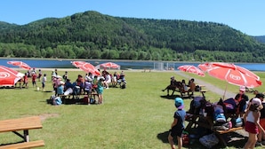 A group of people gathered around picnic tables with red umbrellas, a lake, and forested hills in the background.