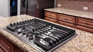 A kitchen with a stainless steel range hood, a built-in gas stove, a stainless steel refrigerator, wooden cabinets, and granite countertops.
