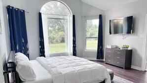 A bedroom with a large bed, a dresser, a TV, and a view of the outdoors.