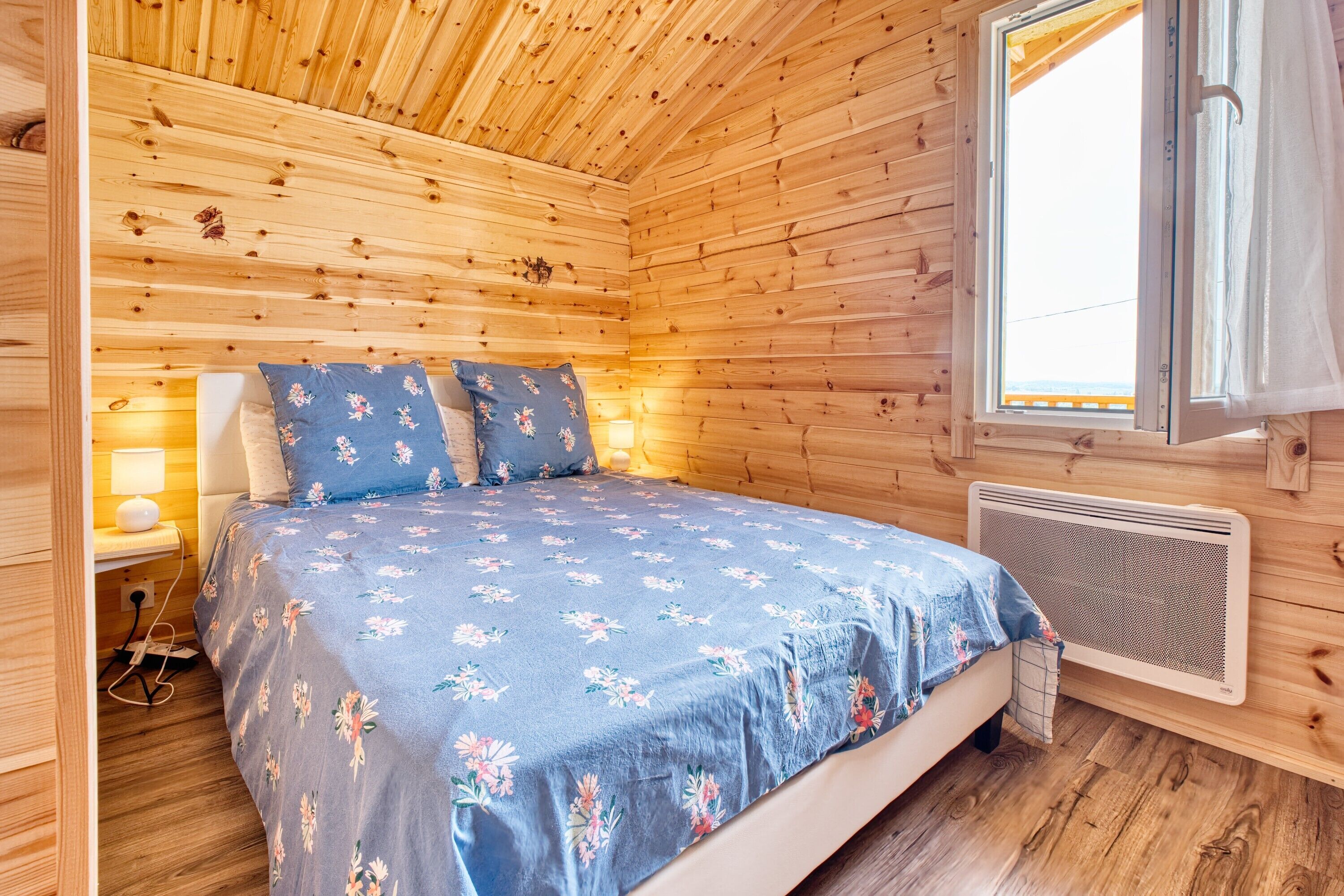 A wooden cabin bedroom with a bed, a nightstand, a lamp, a window with curtains, and a wall-mounted heater.