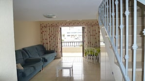 A living room with a sofa, a small table, a balcony with a chair, and a staircase.