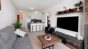 Ein modernes Wohnzimmer mit grauem Sofa, Couchtisch, Fernseher und einer Küche im Hintergrund.