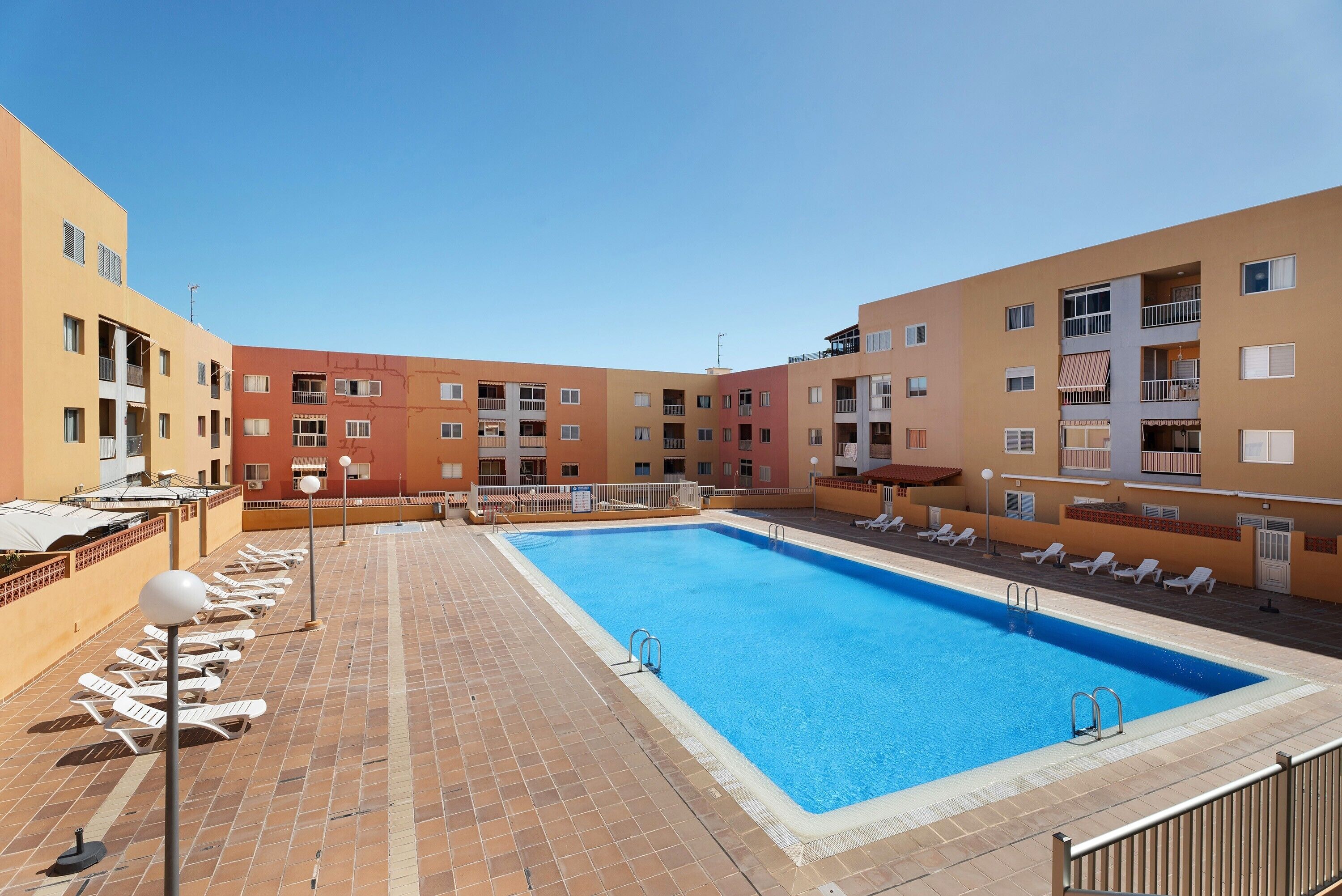 A swimming pool with sun loungers, a tiled deck, and multi-story buildings with balconies.