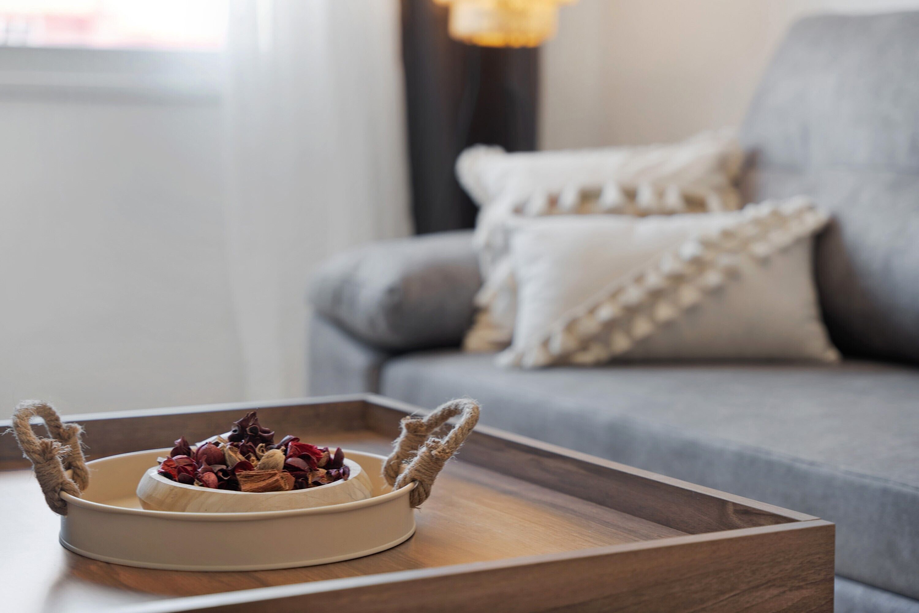 A round, beige ceramic bowl with a rope handle on a wooden tray, placed on a table in front of a grey sofa with white and beige cushions.