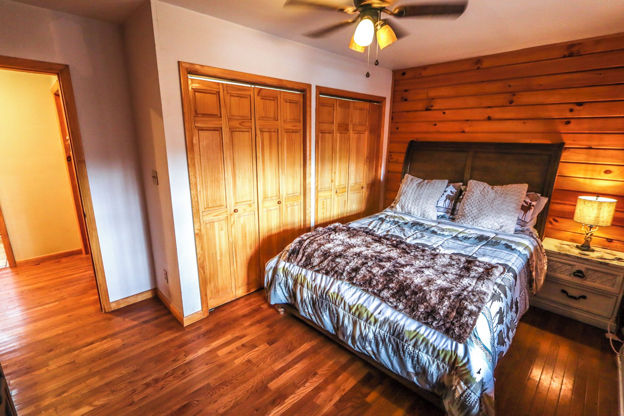 A bedroom with a wooden floor, a bed with patterned bedding, a ceiling fan, and a nightstand with a lamp.