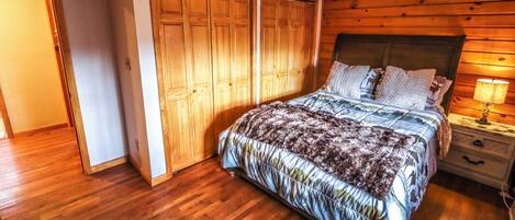 A bedroom with a wooden floor, a bed with patterned bedding, a ceiling fan, and a nightstand with a lamp.