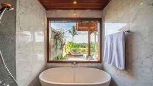 A bathroom with a freestanding bathtub, a shower, and a window with a view of a garden. - Kaki Uma Villa (Sebatu)