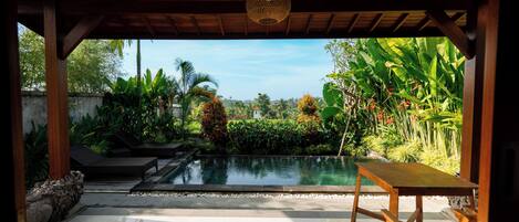 A covered outdoor area with a pool, wooden furniture, and lush tropical plants.