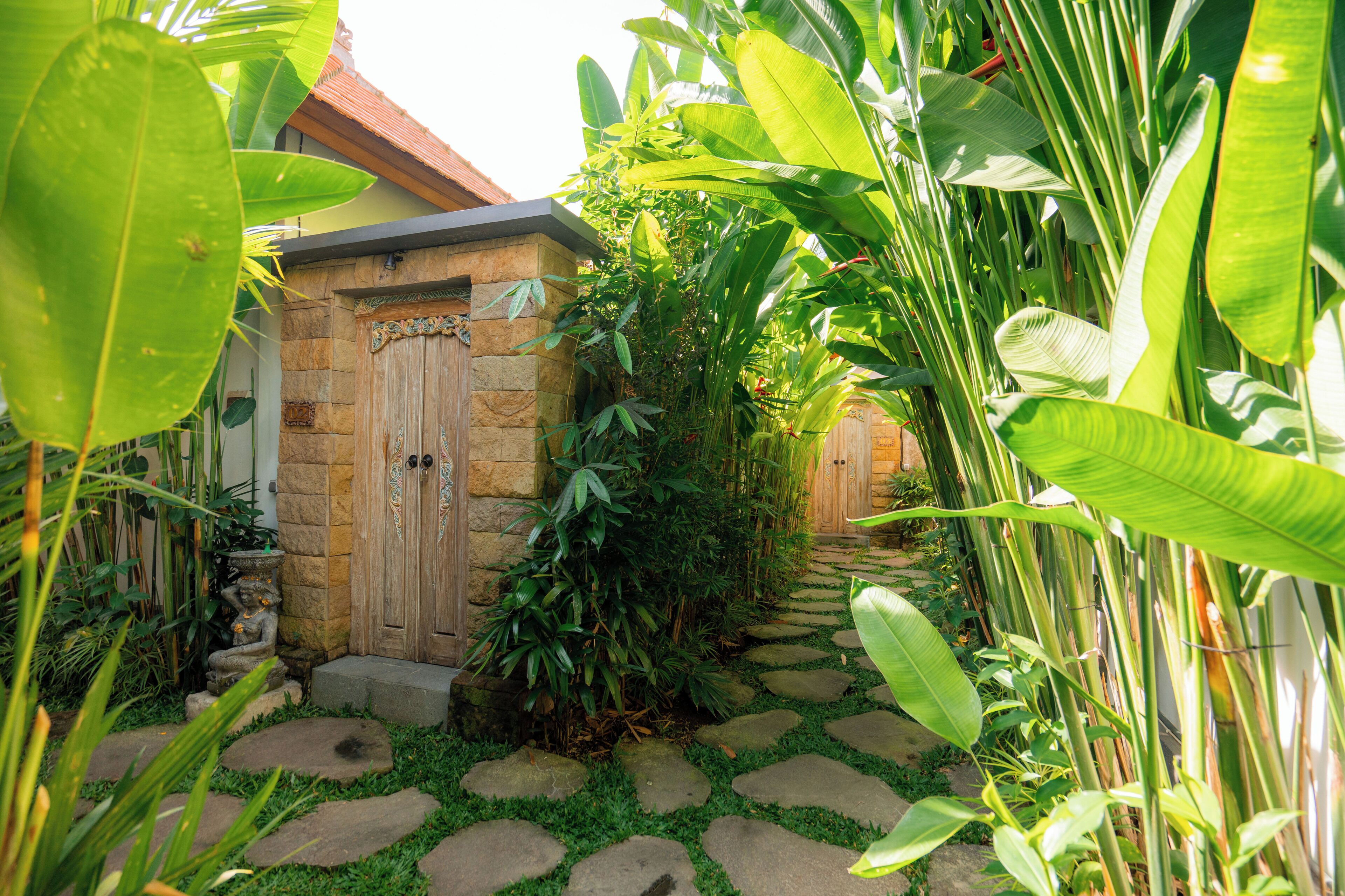 A stone pathway leading to a wooden door, surrounded by lush tropical plants.