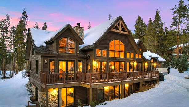 A large, multi-story log cabin with a balcony and large windows, surrounded by snow and trees.