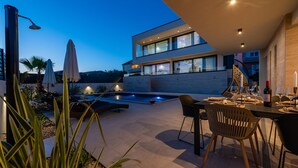 Una zona de menjador a l'aire lliure moderna amb piscina, un edifici de dues plantes i un patio ben il·luminat.