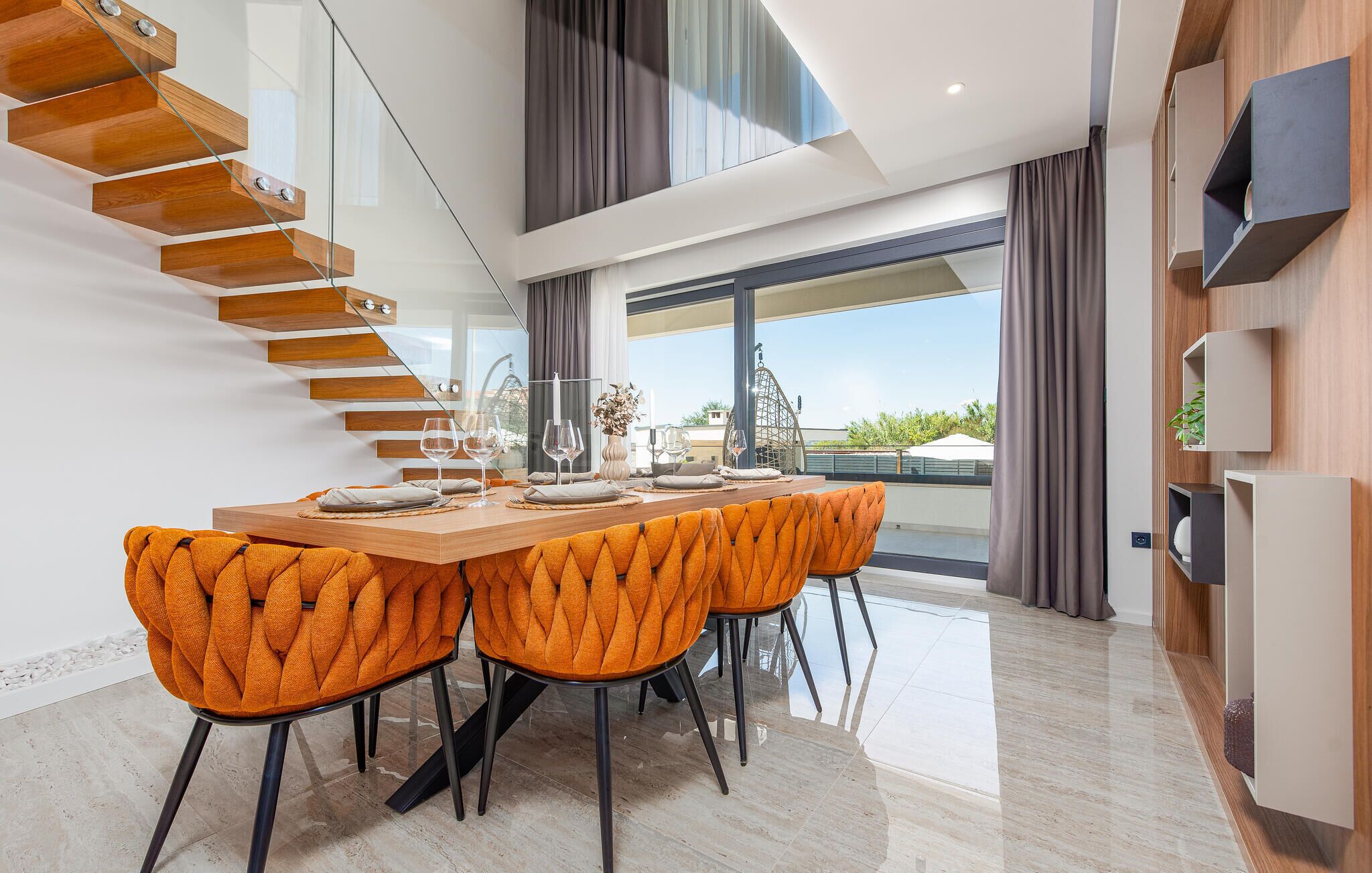 Un comedor moderno con una mesa de vidrio, sillas tapizadas en naranja y una escalera de madera.