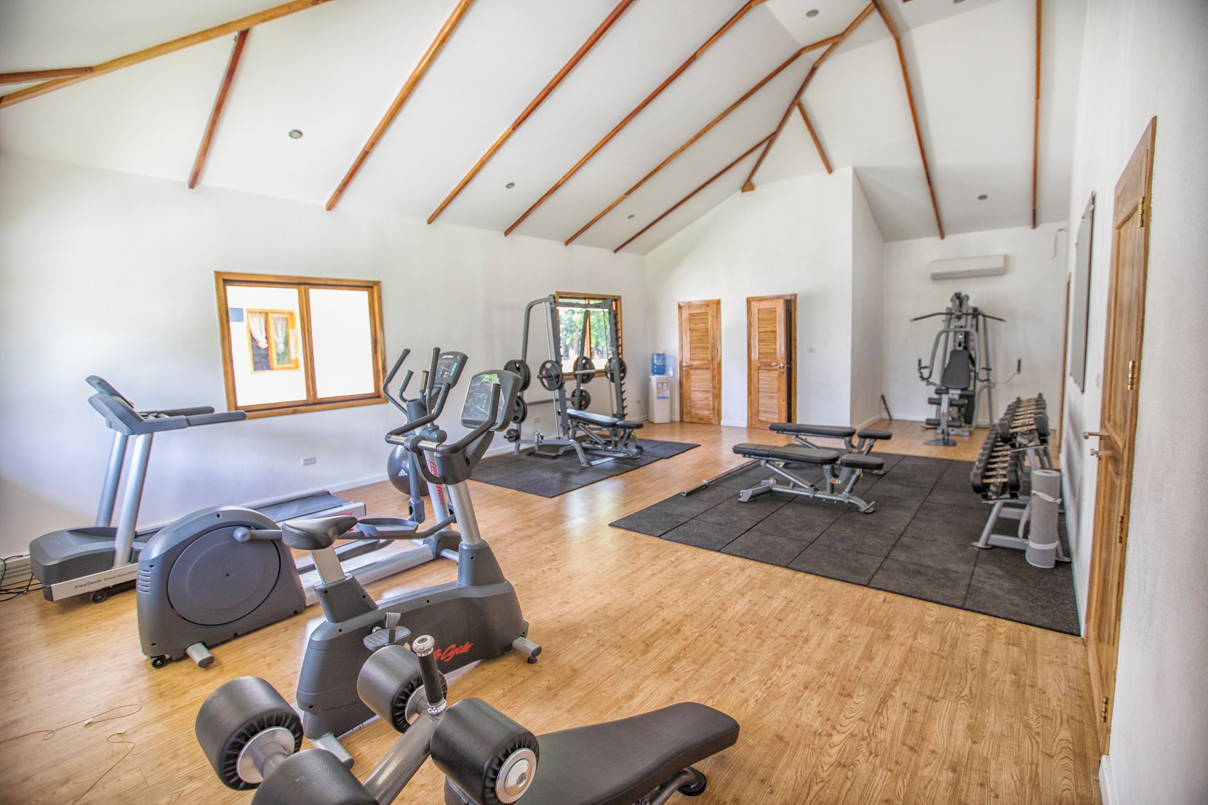 A spacious gym with various exercise equipment, wooden floors, and large windows.