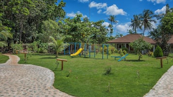 Explore the playground surrounded by lush greenery and palm trees, featuring a slide and climbing structures.