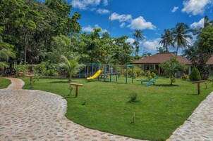 A playground with a yellow slide and blue climbing structures, surrounded by lush greenery and a clear blue sky.