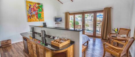 A kitchen with a wooden island, a dining area with wooden chairs, a TV mounted on the wall, and a large window with a view of the beach.