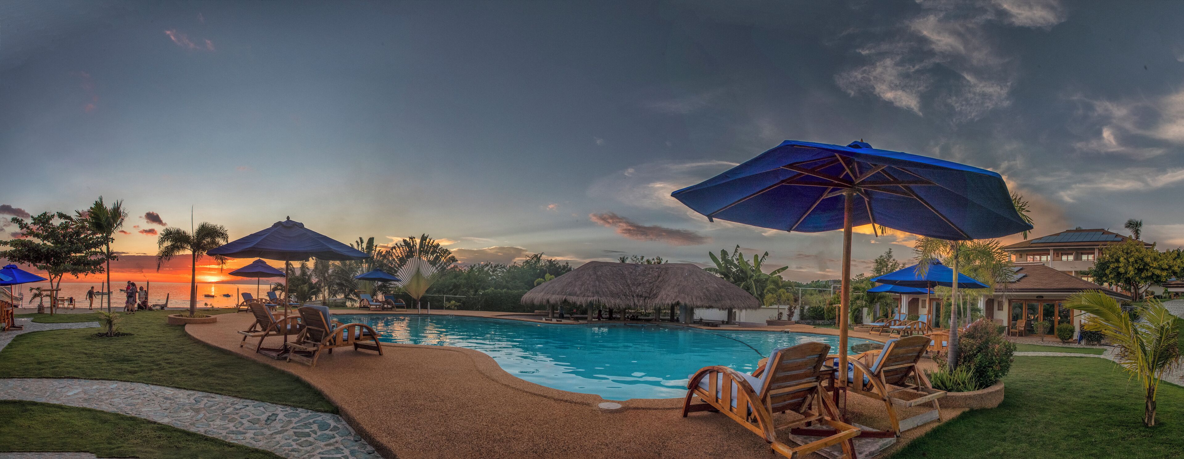 Zona de piscina en un complejo turístico con sillas de descanso y sombrillas, rodeada de palmeras y una estructura con techo de paja.
