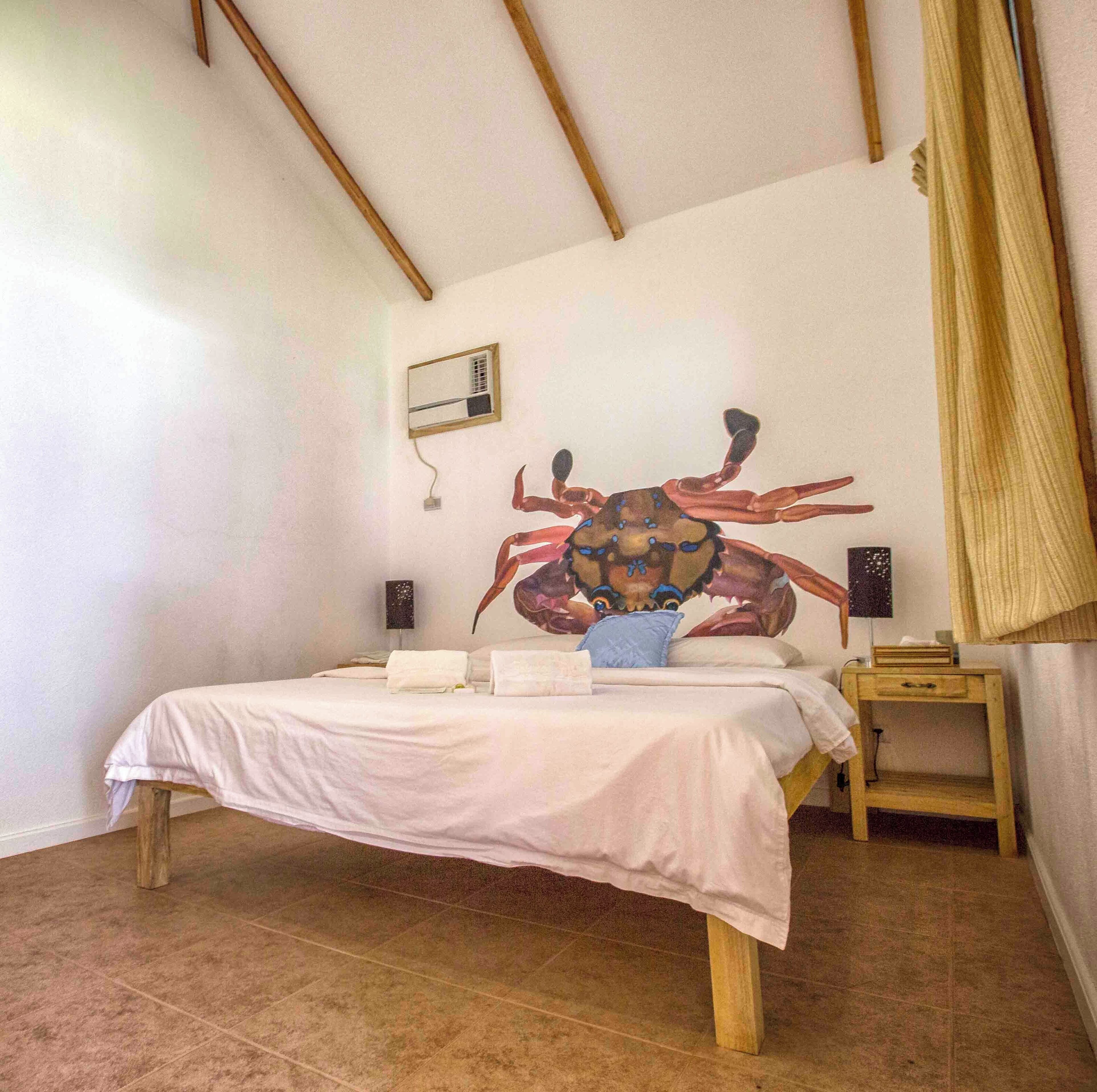 Une chambre à coucher avec un grand lit, une tête de lit en bois, une petite table de chevet, un climatiseur mural et une grande fresque représentant un crabe.