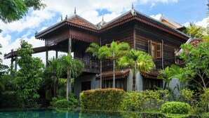 Una casa de madera tradicional con balcón, rodeada de exuberante vegetación y una piscina cristalina.