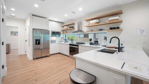 Eine moderne Küche mit weißen Schränken, Holzregalen, einem Edelstahlkühlschrank und einer großen Kochinsel mit Spüle.