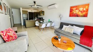 Un salon moderne avec un canapé gris, un plaid rouge et une table basse en bois. On y trouve également un coin repas avec une table et des chaises en verre, et une cuisine à l’arrière-plan.