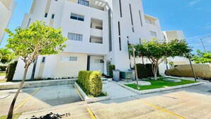 Un immeuble résidentiel moderne à plusieurs étages, doté d’une façade blanche et épurée, d’un petit jardin à l’avant et d’un espace de stationnement.