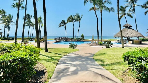 A resort pool area with palm trees, a thatched-roof structure, and a clear view of the ocean. - hermoso depa frente al mar by PartnerHost (Mazatlán)