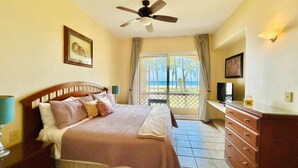 A bedroom with a bed, a dresser, a ceiling fan, a television, and a view of the beach through a window. - hermoso depa frente al mar by PartnerHost (Mazatlán)
