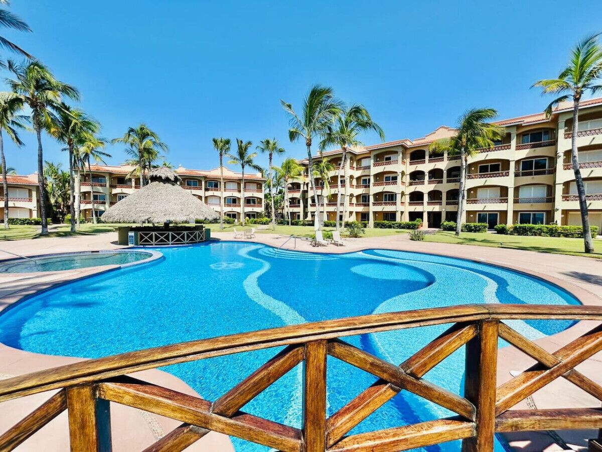 Una piscina di un resort con un pontile di legno, circondata da palme e un edificio a più piani con balconi.