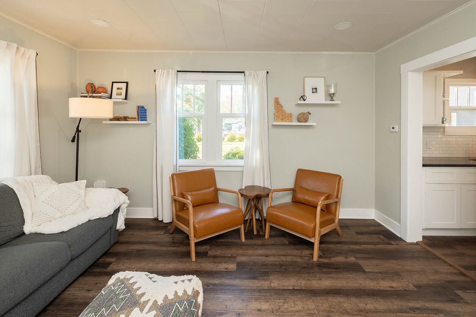 Ein Wohnzimmer mit grauem Sofa, zwei braunen Ledersesseln, einem kleinen Tisch und einem gemusterten Teppich. Ein Fenster mit weißen Vorhängen, eine Lampe und ein Regal mit Dekorationselementen sind ebenfalls zu sehen.