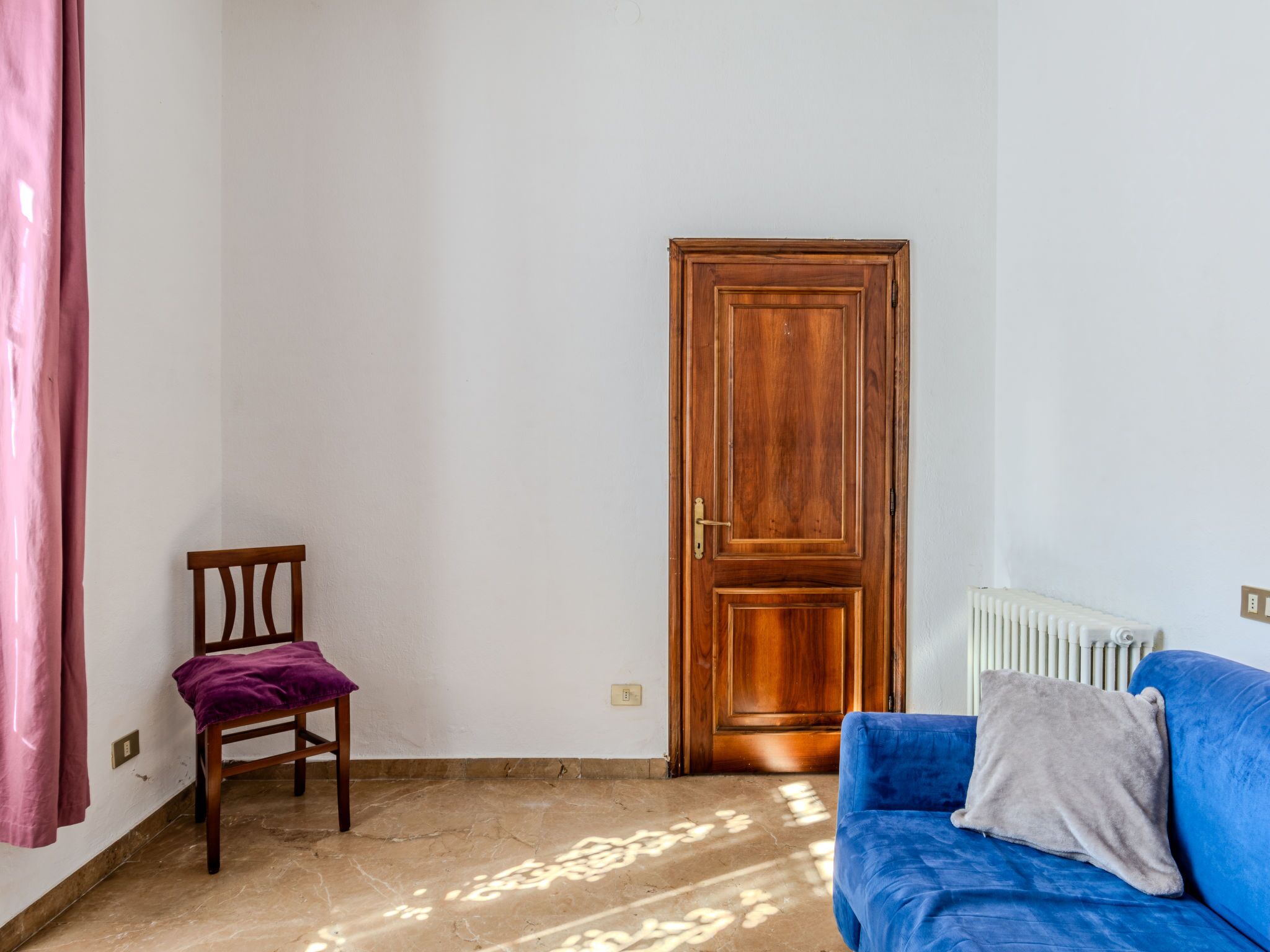 Una habitación con puerta de madera, un sofá azul con almohada gris, una silla de madera con cojín morado y un radiador en la pared.