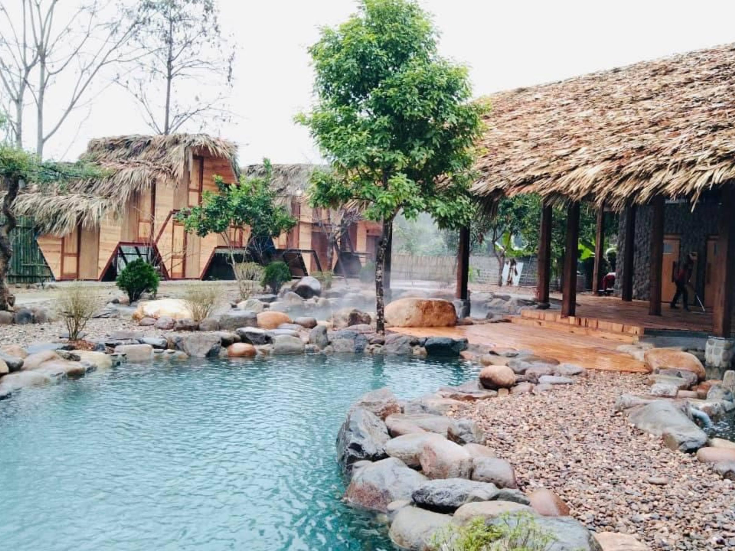 Un espace piscine extérieur paisible, agrémenté d’un aménagement rocheux naturel, entouré de structures à toits de chaume et d’une végétation luxuriante.