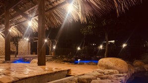 A nighttime outdoor pool area with a thatched roof, stone walls, and illuminated water features. - Dragonfly Hotel Nghia Lo - Free Sauna & Onsen (Nghia Lo)