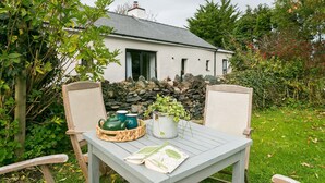 A rustic outdoor seating area with a table and chairs, a stone wall, and a house in the background.