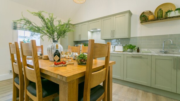 A dining area with a wooden table, six chairs, a vase with greenery, a bottle, and a bowl of salad.