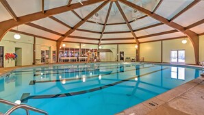 Une piscine intérieure avec des couloirs bien délimités, un plafond haut avec des poutres en bois et une vue sur l’ensemble de l’espace piscine, y compris les vestiaires et une porte donnant sur l’extérieur.