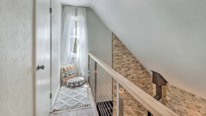 Un escalier étroit avec une rampe blanche, un petit coin salon avec des coussins et une fenêtre ornée de rideaux légers.
