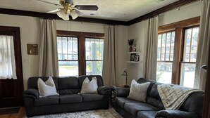 Ein Wohnzimmer mit zwei dunklen Ledersofas, einem gemusterten Teppich, einem Deckenventilator und großen Fenstern mit Vorhängen.