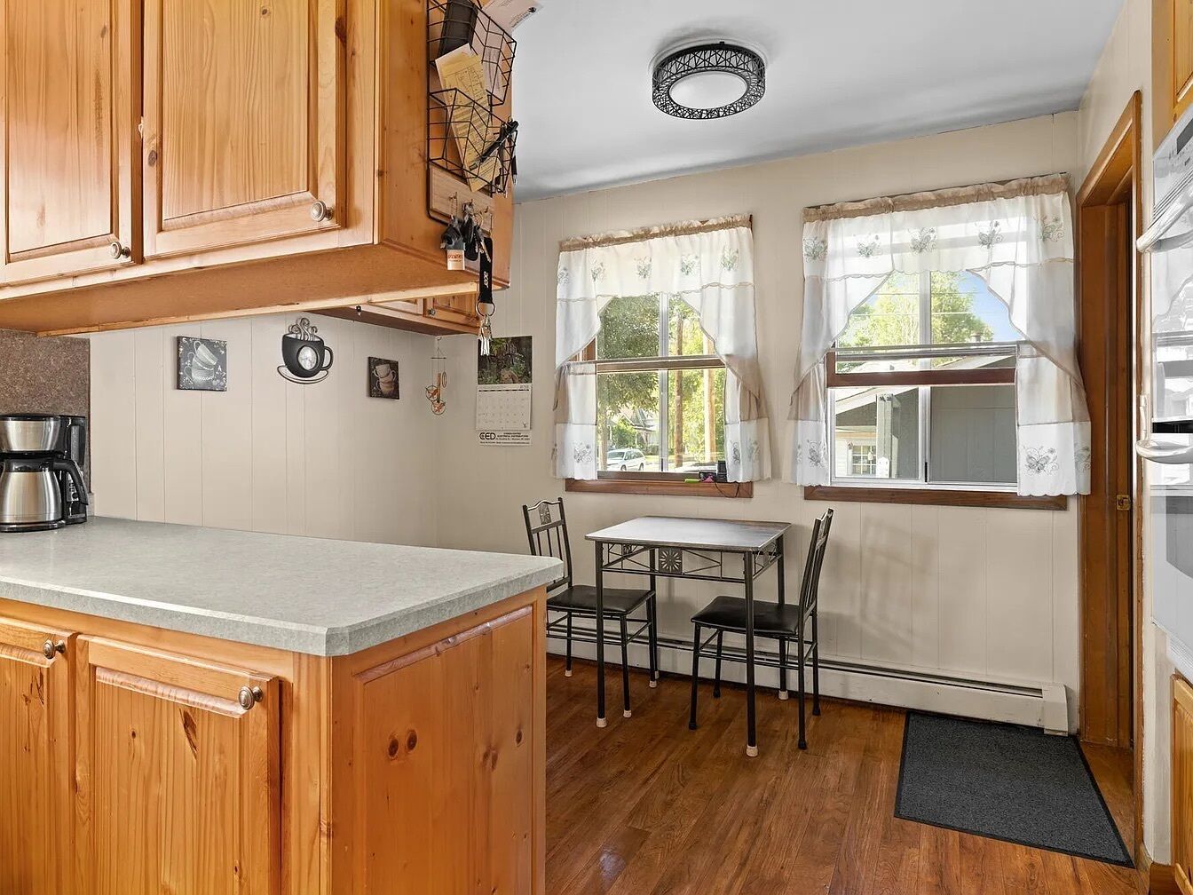 A kitchen with wooden cabinets, a coffee maker, a dining table with chairs, and a window with curtains.