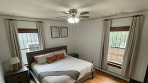 Ein Schlafzimmer mit einem Bett, Nachttischen, einem Deckenventilator und Fenstern mit Vorhängen.