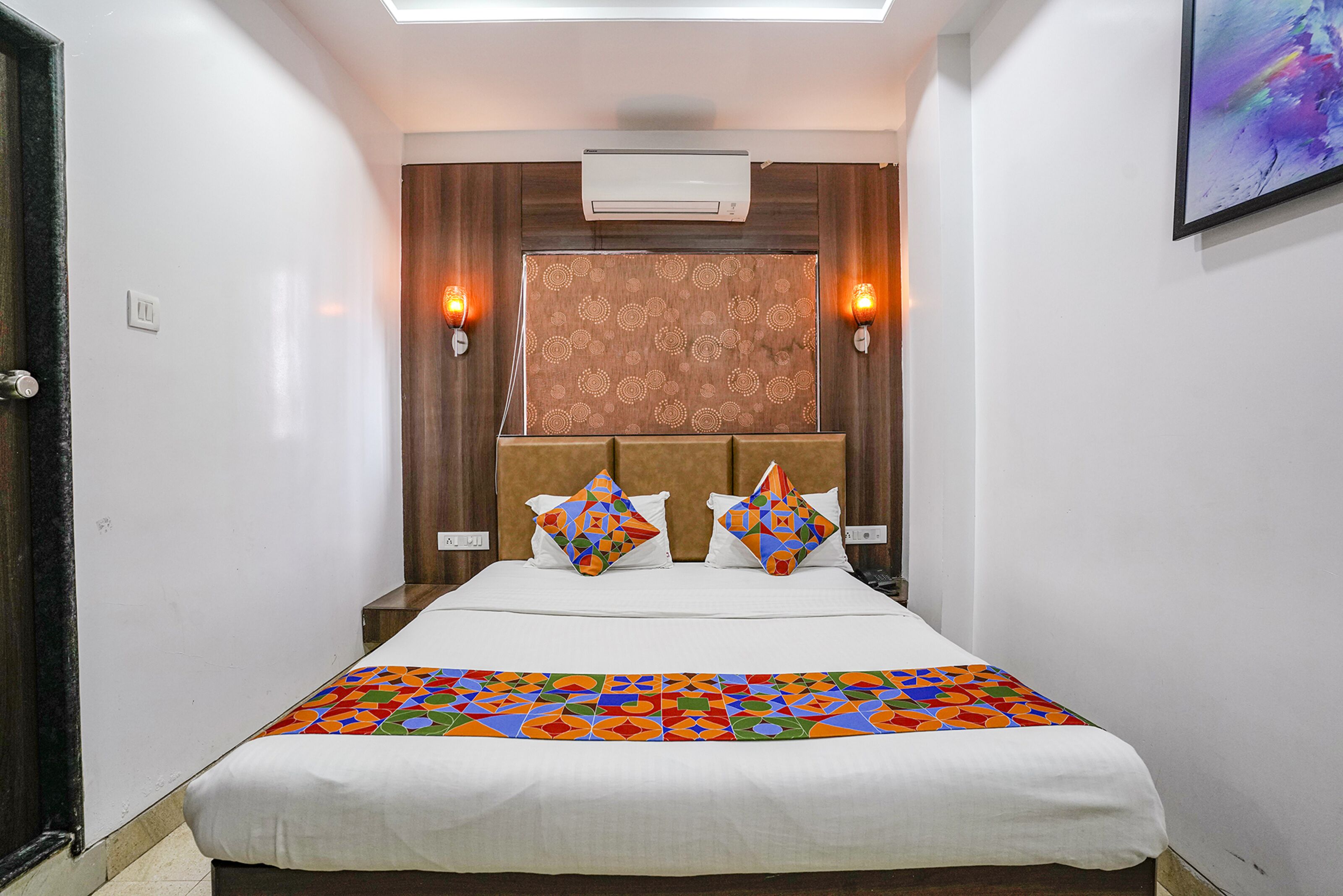 A hotel room with a bed, two wall-mounted lamps, an air conditioner, and a patterned headboard.