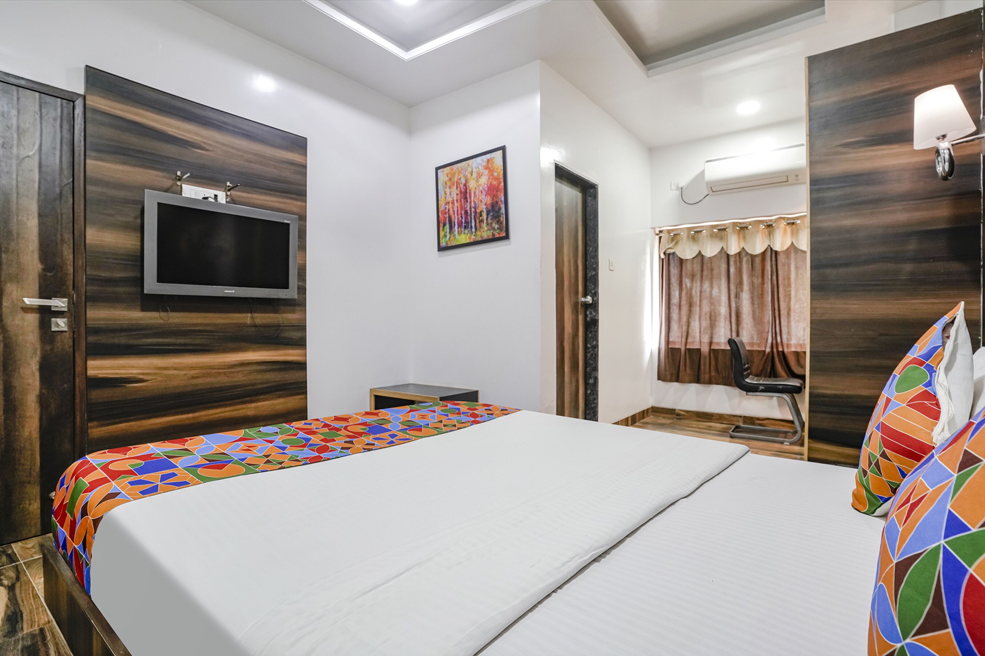 Ein Hotelzimmer mit einem großen Bett, einem an der Wand montierten Fernseher, einem kleinen Tisch und einem Fenster mit Vorhängen.