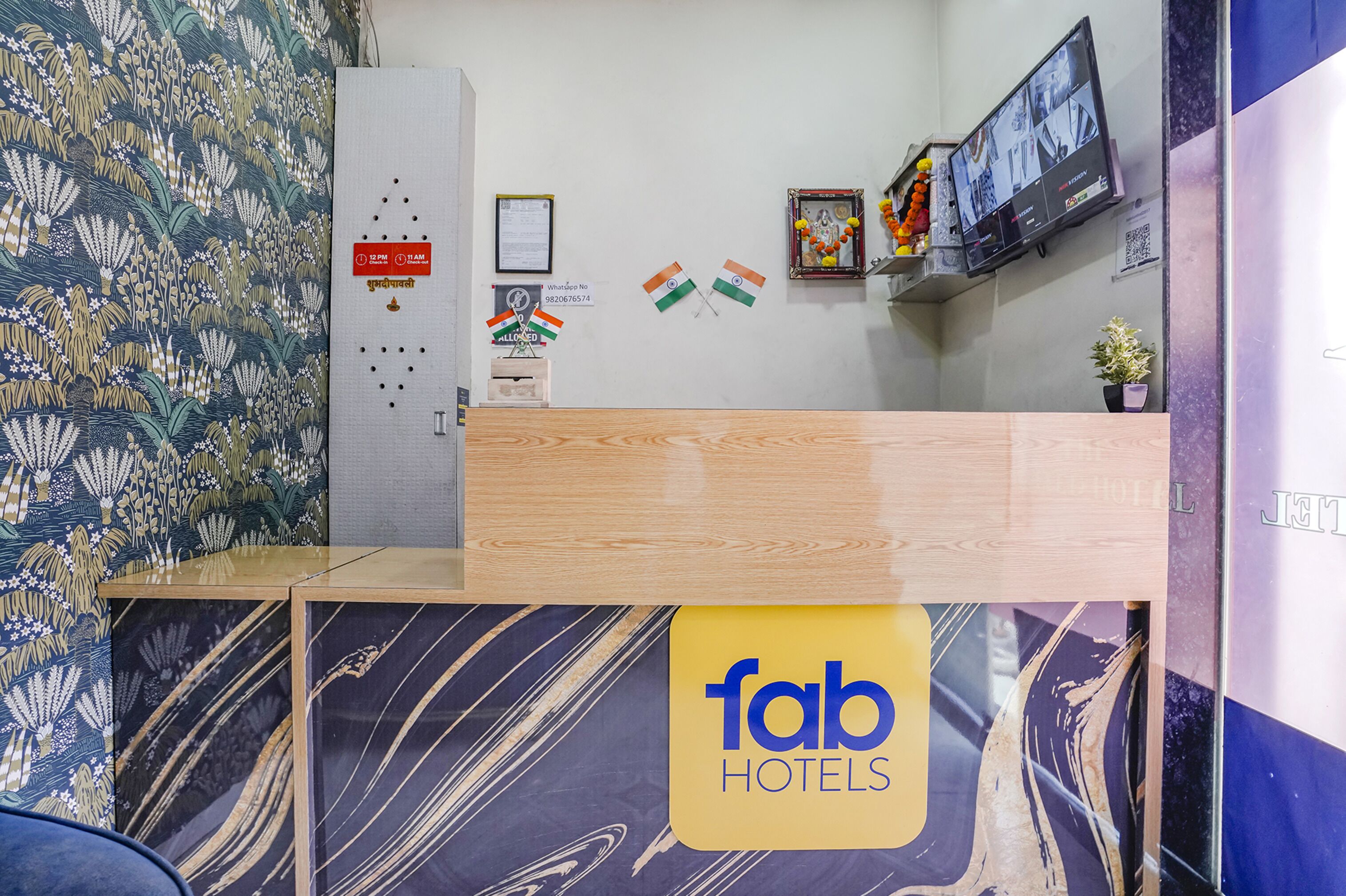 Un comptoir d’accueil d’hôtel avec un comptoir en bois, un mur à motifs de marbre et une enseigne indiquant « fab hotels ». On y voit deux drapeaux, une horloge et un tableau encadré accroché au mur.