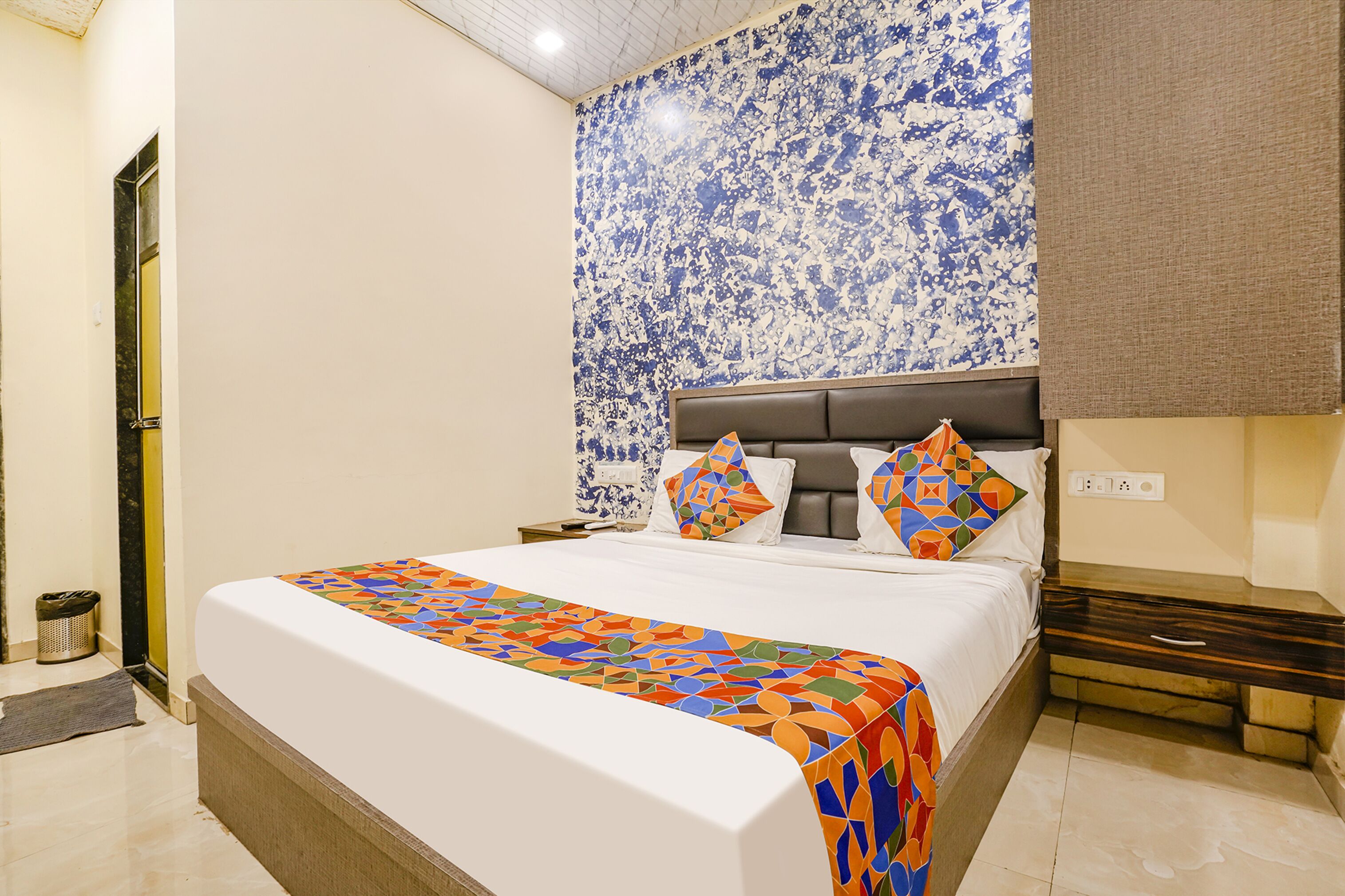 A hotel room with a bed, a nightstand, a wall with a blue and white pattern, and a door.