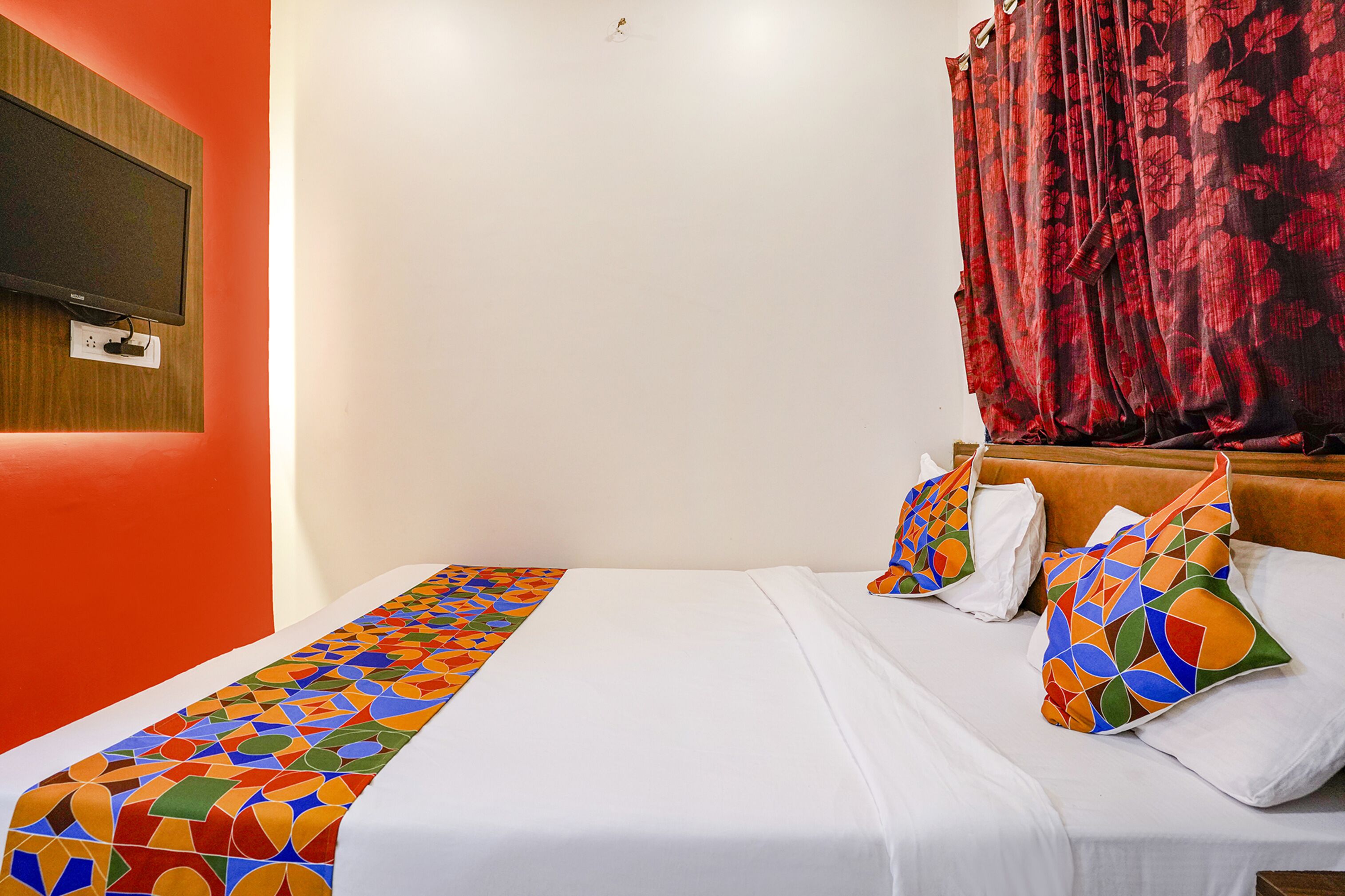 A hotel room with a bed, a TV mounted on the wall, and a window with red curtains.