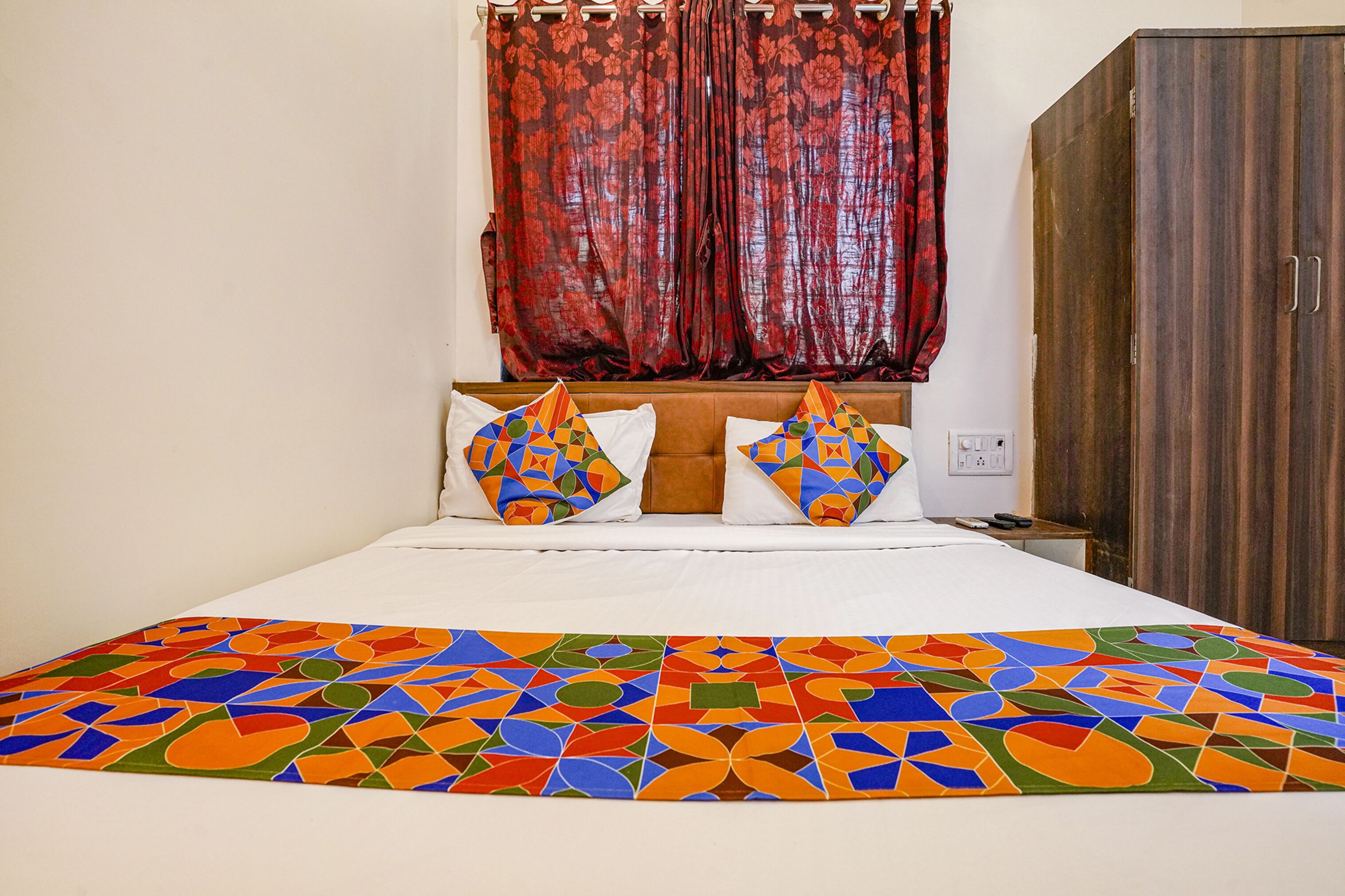 Una cama bien hecha con almohadas y colcha de estampado geométrico colorido, un armario de madera y una ventana con cortinas rojas.
