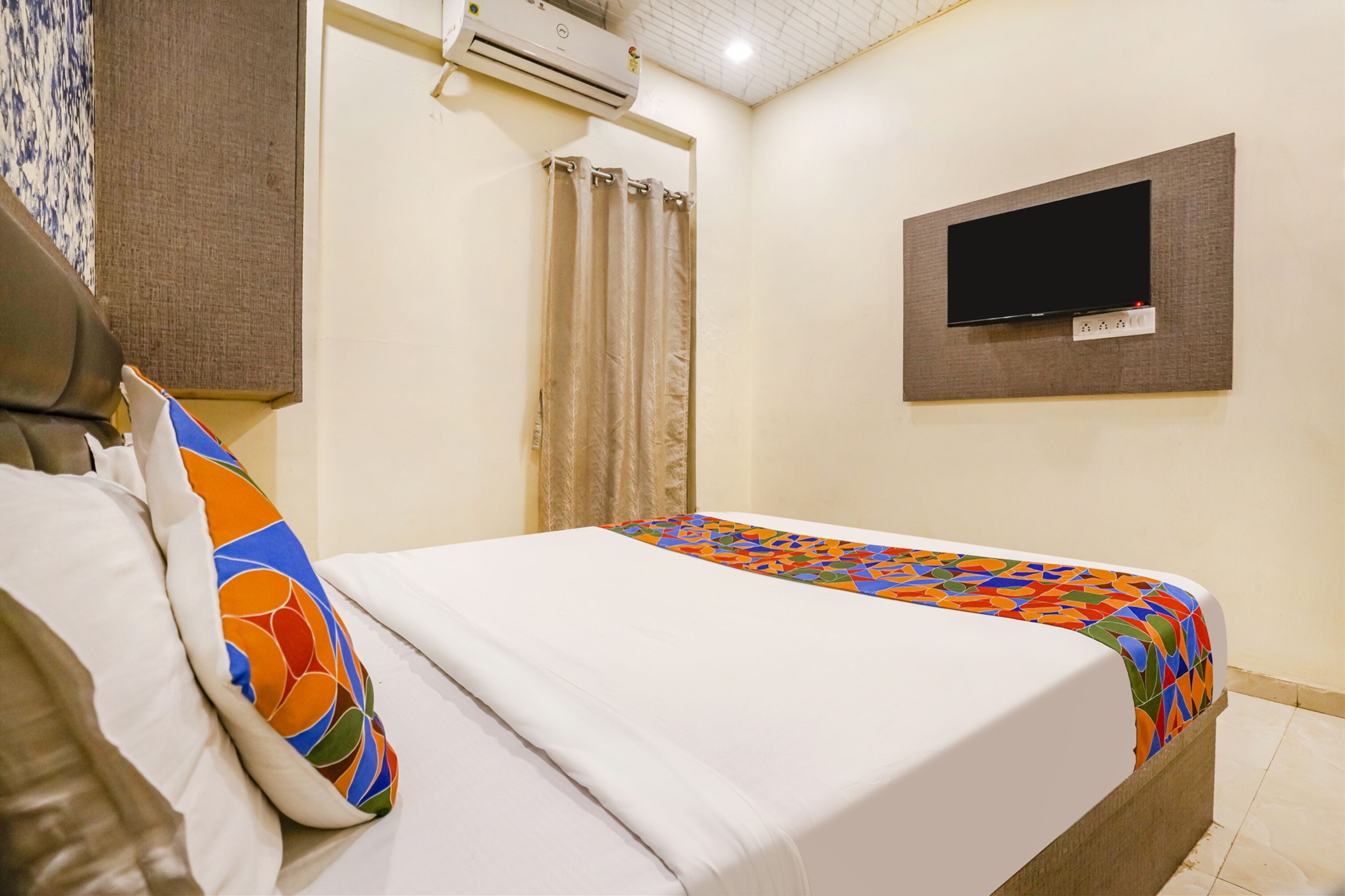 A hotel room with a bed, a TV mounted on the wall, and an air conditioning unit.