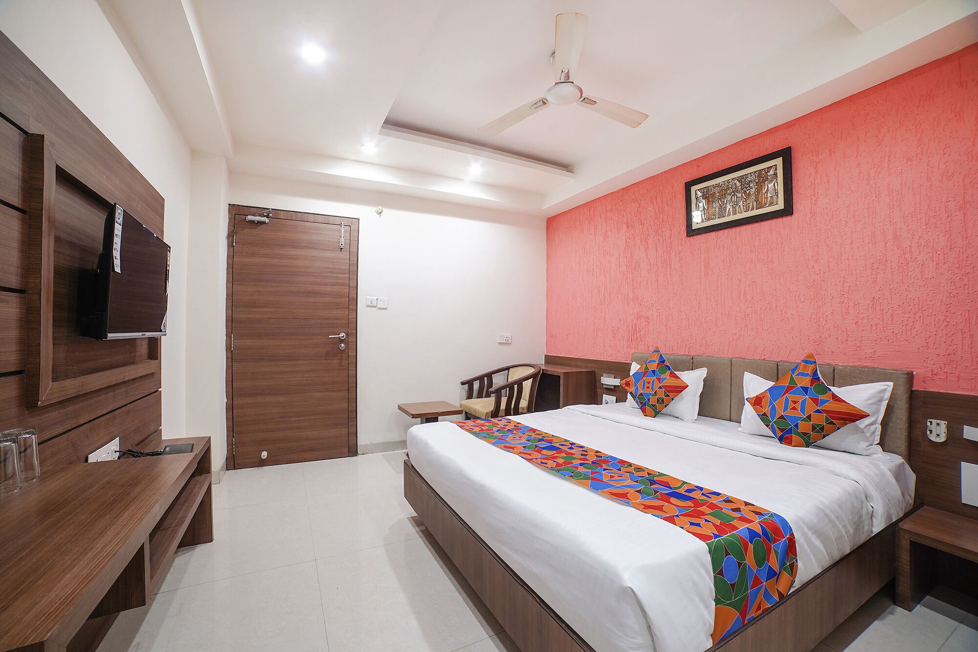 Habitación de hotel con cama, mesitas de noche, televisión, ventilador de techo y una pared de acento roja.
