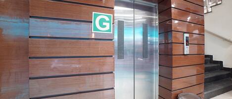 An elevator with a green 'G' sign, a digital display, and a metallic trash can nearby.